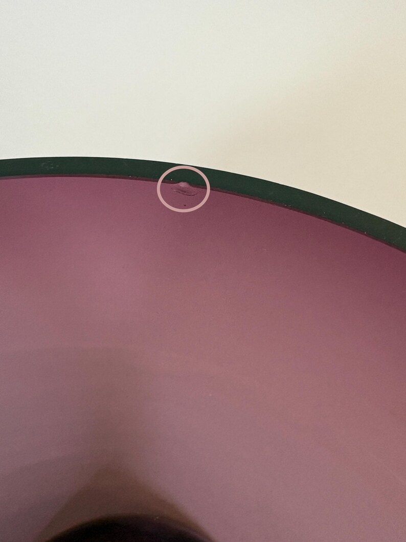 May include: A close-up of a purple glass bowl with a dark green rim. The bowl has a smooth, glossy surface and a slight imperfection on the rim. The background is a plain white.