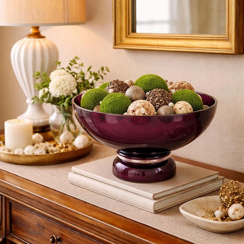 May include: A decorative centerpiece featuring a deep purple bowl filled with green moss balls, decorative spheres, and stones. The bowl sits atop a pedestal and is displayed on a wooden surface, creating an elegant home decor accent.