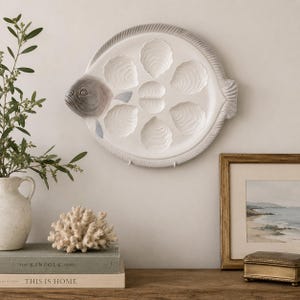May include: A white ceramic serving platter shaped like a flat fish, featuring a grey head and seven molded indentations designed for holding oysters. The platter has a textured fin border and a smooth, glossy finish, displaying a detailed fish face.