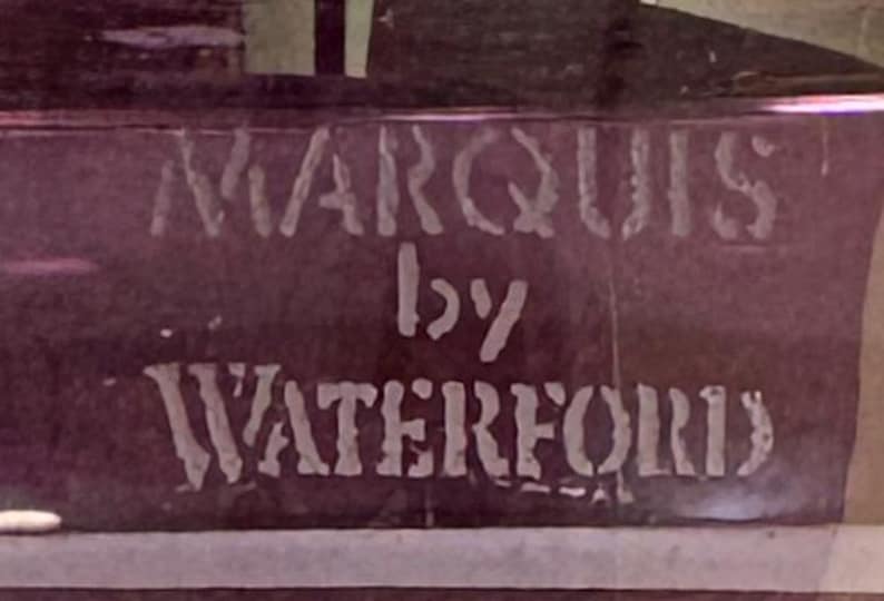 May include: A close-up of a dark purple surface with the words "MARQUIS by WATERFORD" in white, stencil-style lettering. The text is slightly blurred.