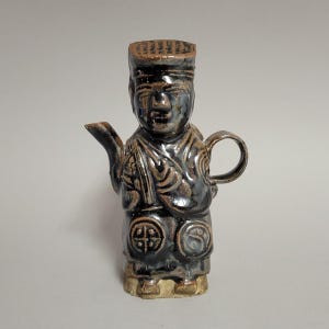 May include: A ceramic teapot shaped like a standing figure with a dark, glossy brown glaze. The figure wears a hat with linear patterns, has a defined face, and features circular embossed designs on the lower garment. It includes a spout and handle.
