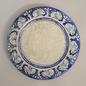 May include: A round ceramic plate with a cream-colored center and a blue border. The border features a repeating pattern of white rabbits and blue floral designs. The plate has a crackle glaze finish.