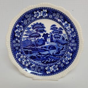May include: A round ceramic plate with a scalloped edge featuring a blue and white transferware pattern. The center depicts a landscape with a stone bridge, a castle, and trees. The border is decorated with floral motifs and small circular patterns.