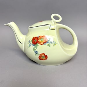 May include: A pale yellow ceramic teapot with a curved handle and spout. The teapot features a floral design of red and orange poppies with blue accents. The lid has a small loop handle. The teapot is a vintage style.