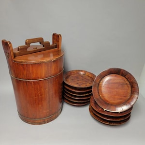 May include: A set of antique wooden serving dishes. The set includes a tall, cylindrical container with a handle and a stack of round plates. The wood has a warm, reddish-brown tone, and the plates have a slightly raised edge.