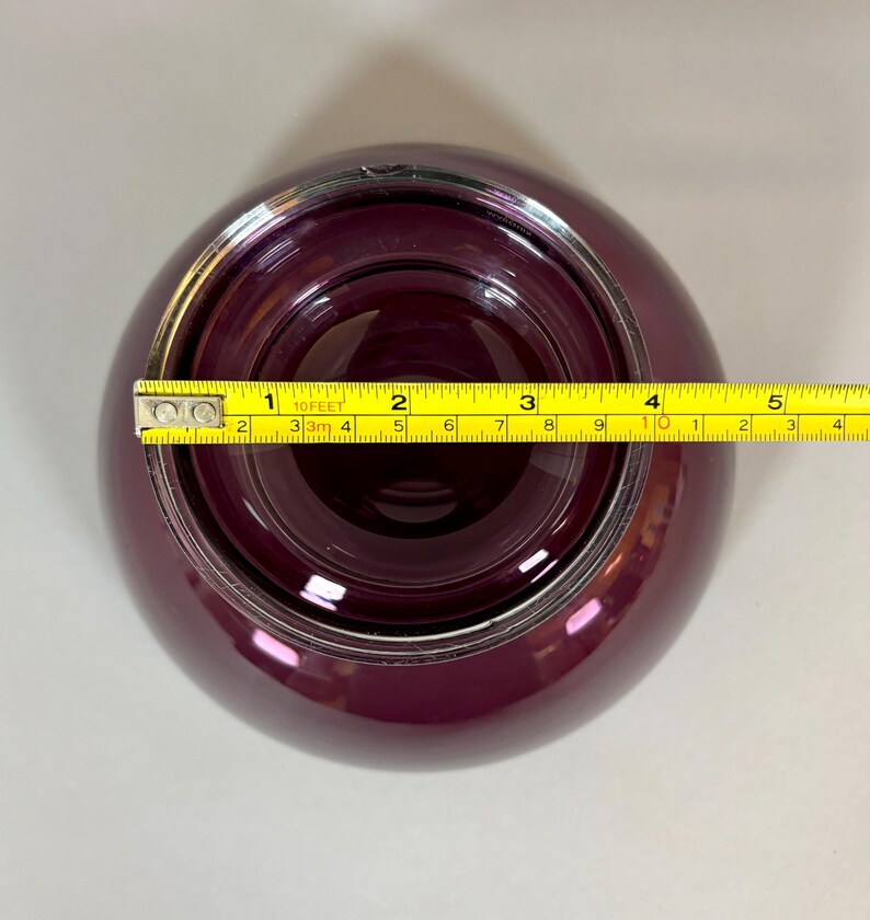 May include: A deep purple glass bowl with a silver rim, viewed from above. A yellow tape measure is placed across the bowl, showing a diameter of approximately 4 inches. The bowl has a rounded shape and a glossy finish.