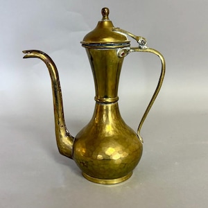 May include: A brass coffee pot with a hammered texture on the bulbous base. The vessel features a long, slender curved spout, a tall neck, and a lid with a small knob handle. A thin, arched handle is attached to the side with a metal hinge.