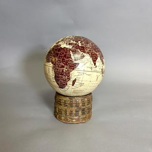 May include: A spherical globe featuring a cream-colored background with dark maroon continents and black text labels for countries and oceans. The globe is mounted on a cylindrical, textured, weathered brown base with a repeating grid pattern.