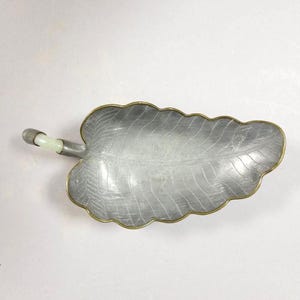 May include: A silver-colored leaf-shaped dish with a brass-colored rim and a jade-colored handle. The dish has detailed veining and a scalloped edge. The handle is cylindrical with a jade-colored stone in the center. The dish is on a white background.