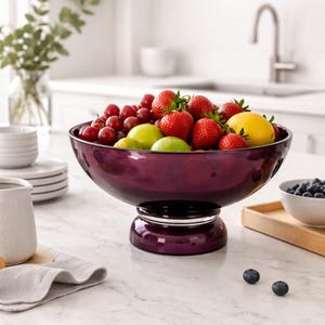 May include: A deep purple glass fruit bowl filled with fresh fruit, including strawberries, cherries, lemons, and limes. The bowl sits on a pedestal base and is displayed on a white countertop. A small bowl of blueberries is in the background.