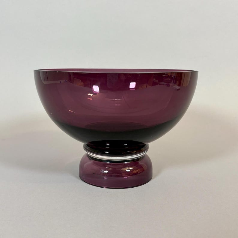 May include: A deep purple glass bowl with a wide, rounded top and a black base. The bowl sits on a short, cylindrical pedestal with a silver band. The glass has a glossy finish.