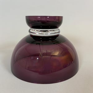 May include: A decorative, deep purple glass bowl with a rounded base and a smaller, cylindrical top. A silver-colored band encircles the bowl's midsection. The bowl is designed with a glossy finish, reflecting light.