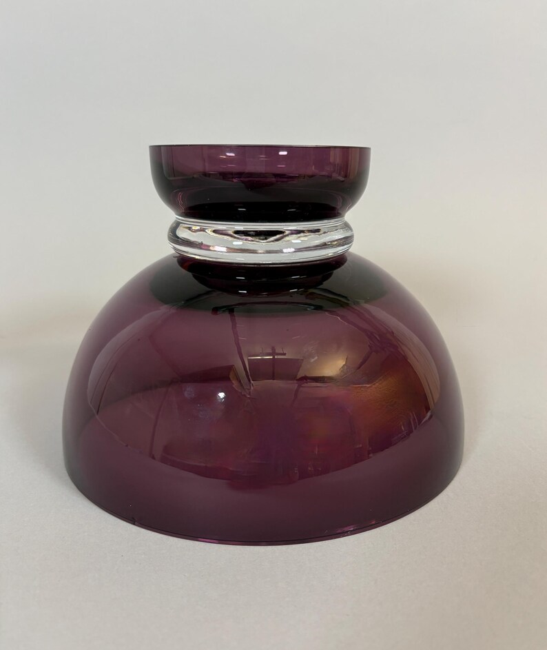 May include: A decorative, deep purple glass bowl with a rounded base and a smaller, cylindrical top. A silver-colored band encircles the bowl's midsection. The bowl is designed with a glossy finish, reflecting light.