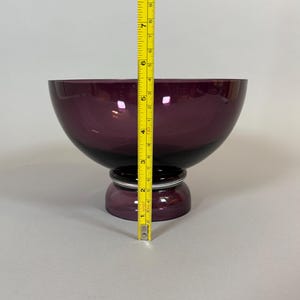 May include: A deep purple glass bowl with a rounded shape, resting on a pedestal base. The bowl measures approximately 7 inches in height, with a dark band at the base of the bowl and a silver accent.