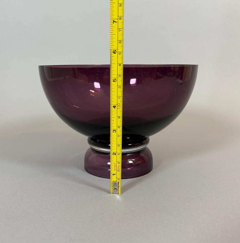 May include: A deep purple glass bowl with a rounded shape, resting on a pedestal base. The bowl measures approximately 7 inches in height, with a dark band at the base of the bowl and a silver accent.