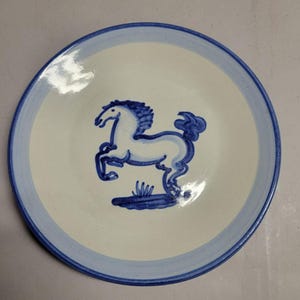 May include: A white ceramic plate with a blue rim and a blue horse illustration in the center. The horse is rearing up on its hind legs. The plate has a light blue border.