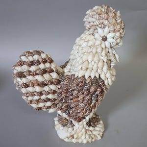 May include: A decorative rooster figurine crafted entirely from small seashells. The body features a mix of white, cream, and dark brown shells arranged in layers to mimic feathers. The head has a crest of shells and a single white flower-like shell eye.