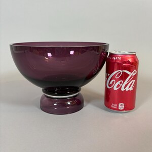 May include: A large, deep purple glass bowl with a pedestal base. The bowl is round with a wide opening and a dark purple base. A red Coca-Cola can is next to the bowl.