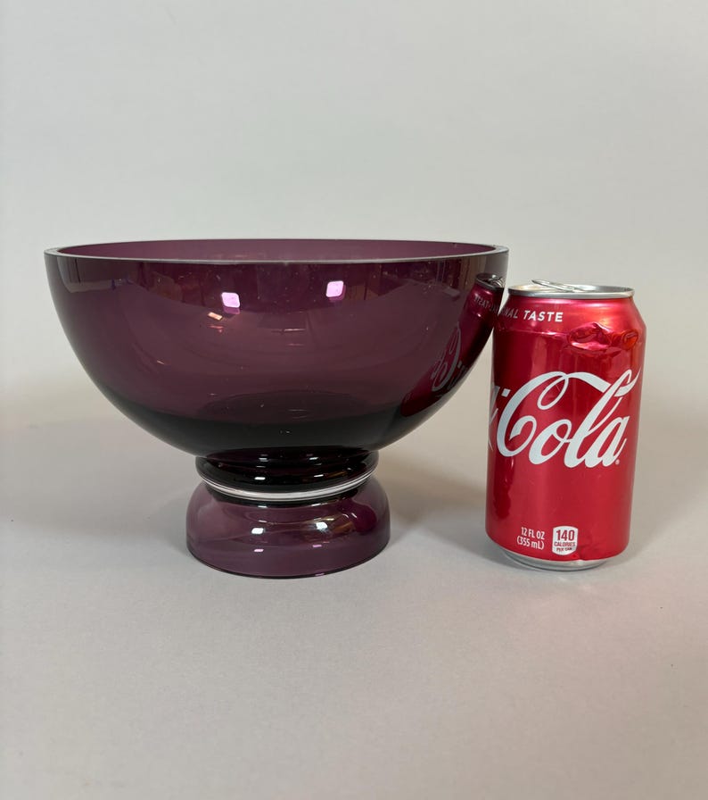 May include: A large, deep purple glass bowl with a pedestal base. The bowl is round with a wide opening and a dark purple base. A red Coca-Cola can is next to the bowl.