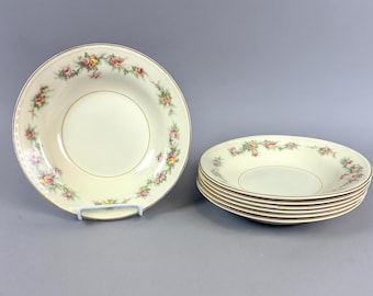 May include: A set of cream-colored bowls with a delicate floral pattern in pink, yellow, and green. The bowls have a gold rim and are stacked neatly. One bowl is displayed on a white stand.
