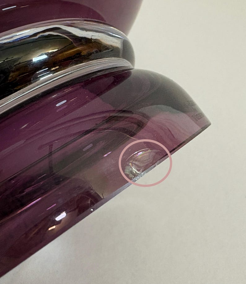 May include: Close-up of a purple glass vase with a silver band. The vase has a rounded base and a flared top. The glass has a glossy finish and a slight imperfection on the edge.
