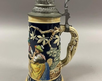 May include: A ceramic beer stein with a detailed relief design. The stein features a dark blue background with a scene of people, a decorative handle, and a metal lid. The base reads "LANG GENUSS".