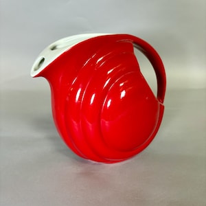 May include: A bright red ceramic pitcher with a glossy finish and a white interior. The pitcher features a rounded, spherical body with concentric circular ridges on the exterior and a curved handle extending from the top to the side.