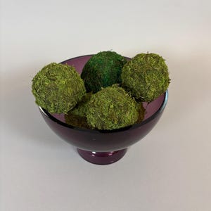May include: A deep purple glass bowl filled with five moss-covered spheres. The spheres vary in shades of green, creating a natural, textured appearance. The bowl has a pedestal base, adding to its decorative appeal. Ideal for home decor.