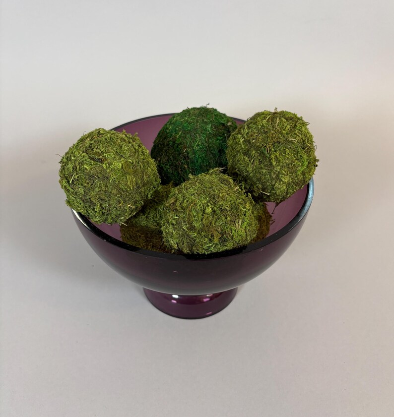 May include: A deep purple glass bowl filled with five moss-covered spheres. The spheres vary in shades of green, creating a natural, textured appearance. The bowl has a pedestal base, adding to its decorative appeal. Ideal for home decor.