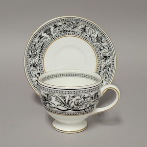 May include: A white teacup and saucer set with black ornate designs and gold trim. The teacup has a pedestal base and a curved handle. The saucer is round and complements the teacup's design. The set is likely porcelain.
