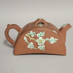 May include: A brown ceramic teapot with a curved handle and spout. The teapot is decorated with green and white floral designs. The lid has a small handle and is also decorated with flowers. The teapot is likely used for brewing tea.