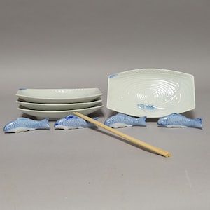May include: A white ceramic sushi set with blue fish-shaped chopstick rests. The set includes rectangular plates and a large serving platter with a fish design. A pair of wooden chopsticks rests on the table.