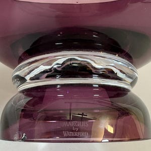 May include: A close-up of a Marquis by Waterford purple glass bowl. The bowl has a wide, rounded top and a clear glass band around the middle. The base is a darker shade of purple. The brand name is visible on the base.
