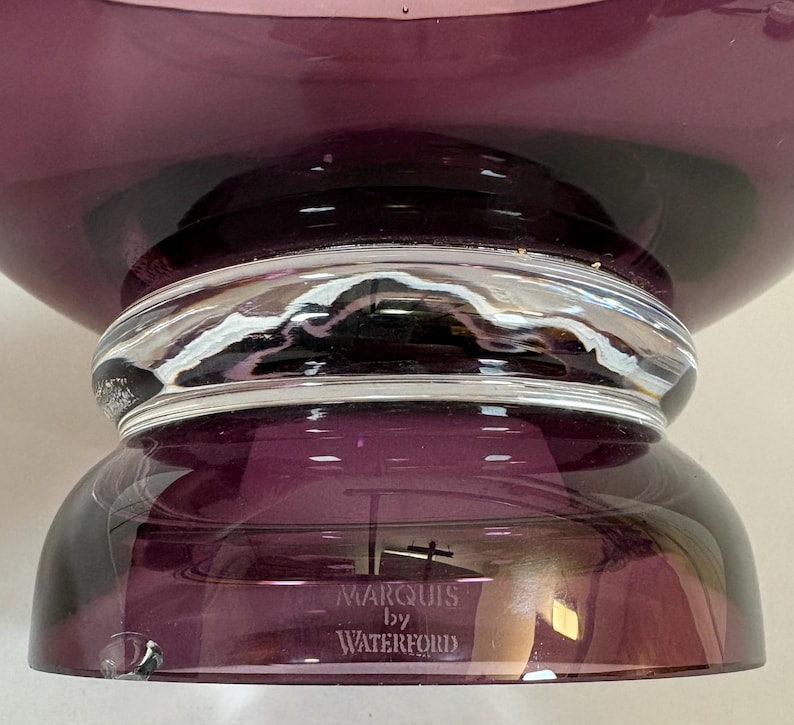 May include: A close-up of a Marquis by Waterford purple glass bowl. The bowl has a wide, rounded top and a clear glass band around the middle. The base is a darker shade of purple. The brand name is visible on the base.