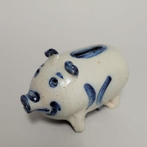 Puede incluir: Una alcancía de cerámica con forma de cerdo. El cuerpo blanco está decorado con detalles azules en las orejas, los ojos, el hocico y los lados. Tiene una ranura para monedas en la parte superior.