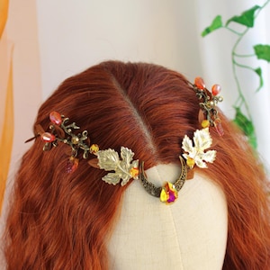 Puede incluir: Una corona dorada con temática de hojas, con piedras preciosas ámbar y amarillas. La tiara presenta hojas de metal detalladas y pequeñas cuentas de colores, creando un diseño elegante inspirado en la naturaleza. La corona se exhibe en una cabeza de maniquí con cabello largo y castaño rojizo.