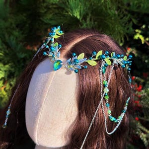 May include: A handcrafted headpiece featuring a teal and green color scheme. The design includes a central jewel, beaded accents, and delicate leaf details. Strands of beads cascade down, adding a flowing element to the design. The headpiece is set against a backdrop of green foliage.