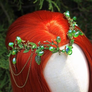 May include: A green and gold-toned headpiece with leaf and crystal accents. The headpiece features a delicate design with green crystals and small leaves. The headpiece is worn on a mannequin head with red hair, set against a green background.