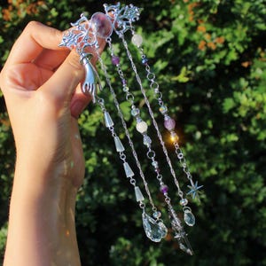 May include: Silver hair accessory with a filigree design, a purple gemstone, and dangling chains adorned with beads and crystals. The accessory includes a small hand charm and a star-shaped charm.