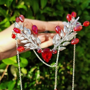 Peut inclure: Peigne argenté avec des accents de cristal rouge et des chaînes pendantes. Le peigne présente un motif de feuilles avec un grand cristal rouge au centre.