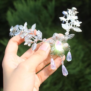May include: A silver-toned metal headpiece featuring intricate leaf and floral filigree patterns. The design is accented with clear crystals and iridescent, teardrop-shaped dangles that hang from the central flower element.