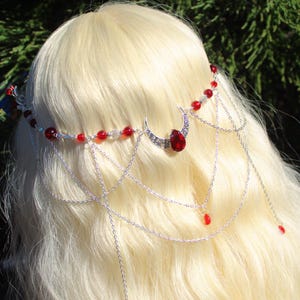 May include: A silver-toned headpiece with a crescent moon design and a large red gemstone. The headpiece features a series of delicate chains with red bead accents and smaller red teardrop-shaped gems. The headpiece is worn on blonde hair.