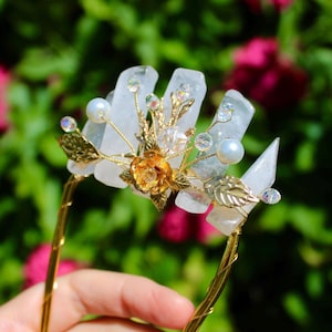 May include: A handcrafted hair accessory featuring clear quartz crystals, faux pearls, and gold-toned metal accents. The design includes a gold flower and delicate leaf details, creating an elegant and natural aesthetic. The hairpiece is held in front of a blurred green and pink background.