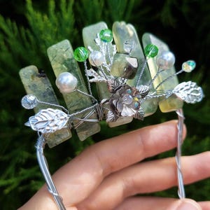 Op de afbeelding: Een zilveren draad haar kam met groene en heldere kristallen, witte parels en een zilveren bloemenaccent.