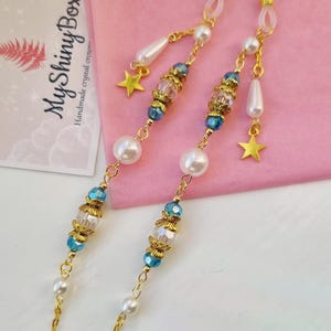 Könnte beinhalten: Goldene Ketten-Brillenhalterung mit blauen und klaren Kristallperlen, weißen Perlen und goldenen Sternanhängern. Die Kette ist an durchsichtigen Kunststoffschlaufen befestigt.