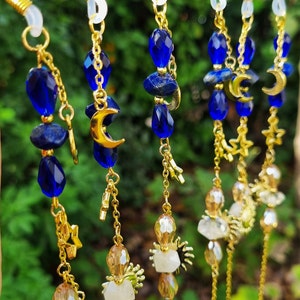Könnte beinhalten: Fünf goldene Brillenketten mit blauen und klaren Glasperlen, goldenen Halbmond-Anhängern und goldenen Stern-Anhängern. Die Ketten hängen an einem Ast.