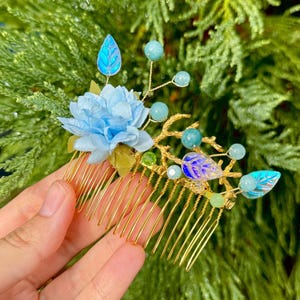 Puede incluir: Un peine para el cabello dorado con una flor de tela azul claro, cuentas azules y verdes y hojas decorativas. El peine tiene una serie de púas finas y paralelas. El fondo es un follaje verde borroso.