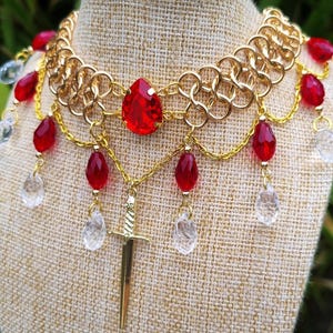 May include: Gold chainmail choker necklace with a large red gemstone pendant and a gold sword charm. The necklace is adorned with red and clear teardrop-shaped beads.