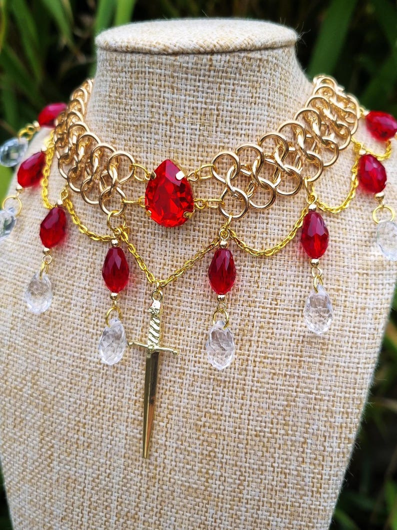 May include: Gold chainmail choker necklace with a large red gemstone pendant and a gold sword charm. The necklace is adorned with red and clear teardrop-shaped beads.