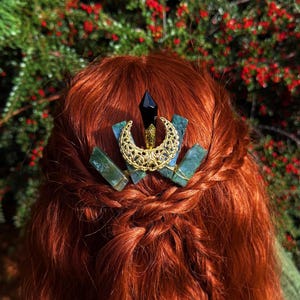 Puede incluir: Un accesorio para el cabello de color dorado con un diseño de media luna, una gema negra y detalles de piedras verdes. El adorno para el cabello se coloca sobre un fondo de cabello rojo y un fondo borroso de bayas rojas y follaje verde.